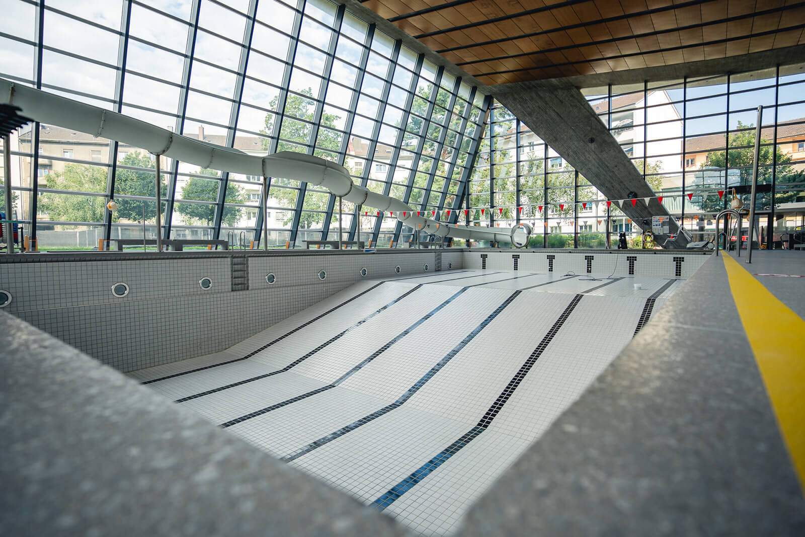 Ein leeres Hallenbad mit gefliestem Boden und markierten Schwimmbahnen, umgeben von grossen Glasfenstern und einer Holzdecke.