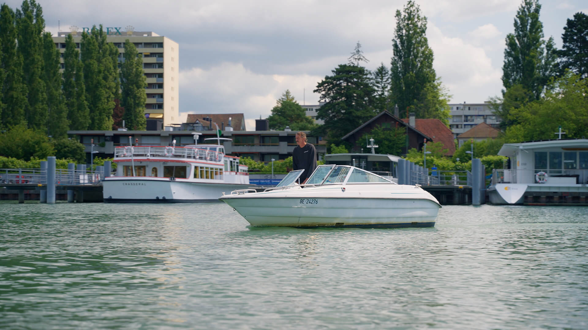 Eine Person steht auf einem kleinen weißen Motorboot in einem ruhigen Gewässer, im Hintergrund sind eine angedockte Fähre und Gebäude zu sehen - Szenen aus dem Alltag vom Leiter Eismeister in der Tissot Arena.