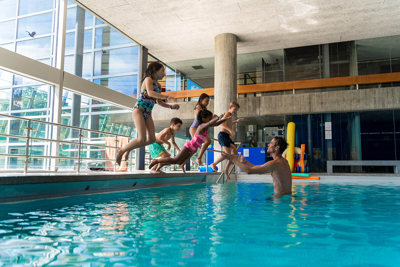 Ein Mann in einem Schwimmbecken streckt seine Hand aus, als vier Kinder vom Beckenrand eines Hallenbades auf ihn zu springen.