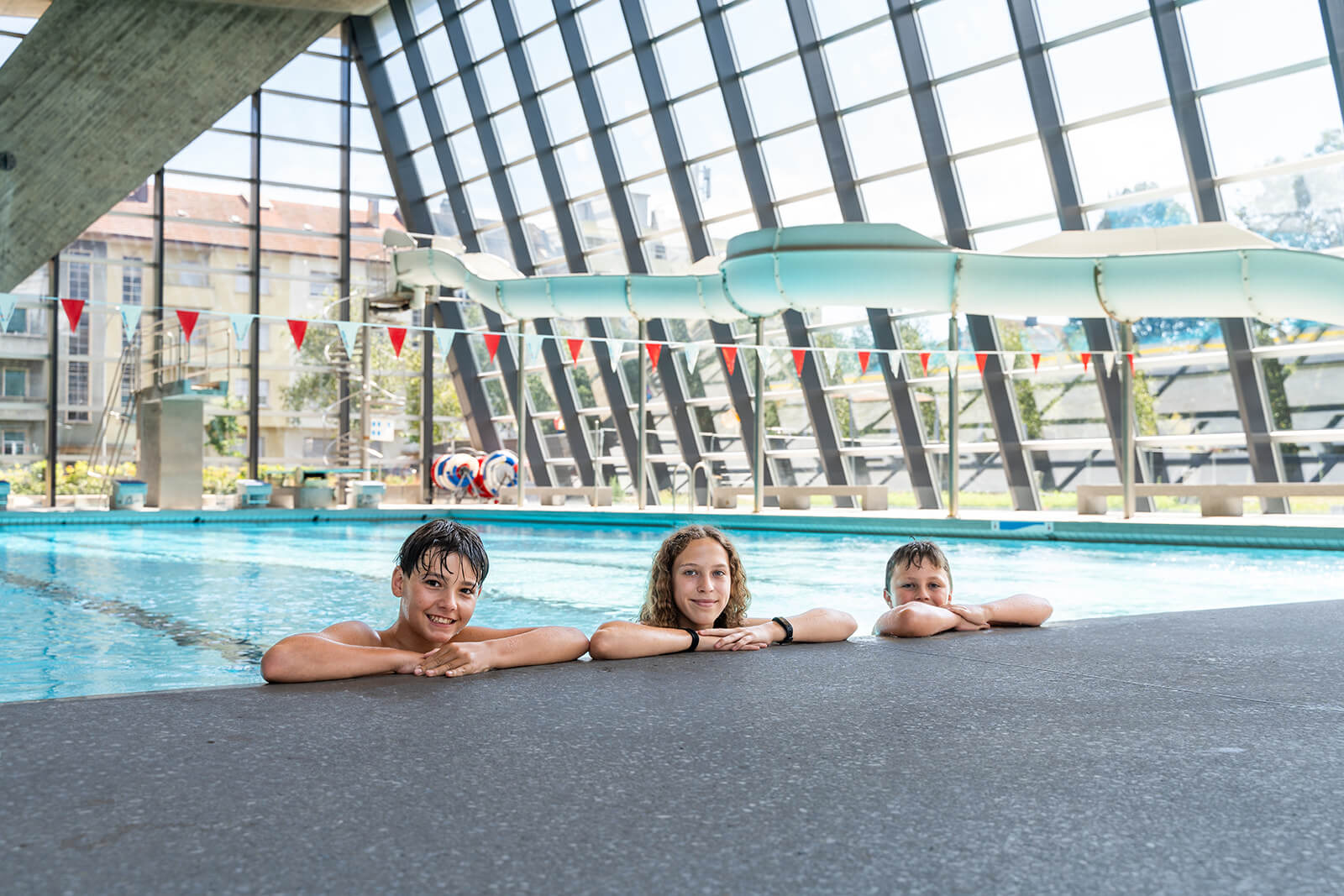 Drei Kinder lächeln und ruhen sich am Rande eines Hallenbades aus, mit einer Wasserrutsche und großen Glasfenstern im Hintergrund.