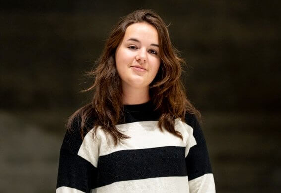 Eine junge Frau mit langen braunen Haaren und einem schwarz-weiß gestreiften Pullover steht vor einem schlichten, dunklen Hintergrund. Ihr Gesichtsausdruck spiegelt die ruhige Konzentration wider, die in Der Alltag vom Leiter Eismeister in der Tissot Arena zu sehen ist.