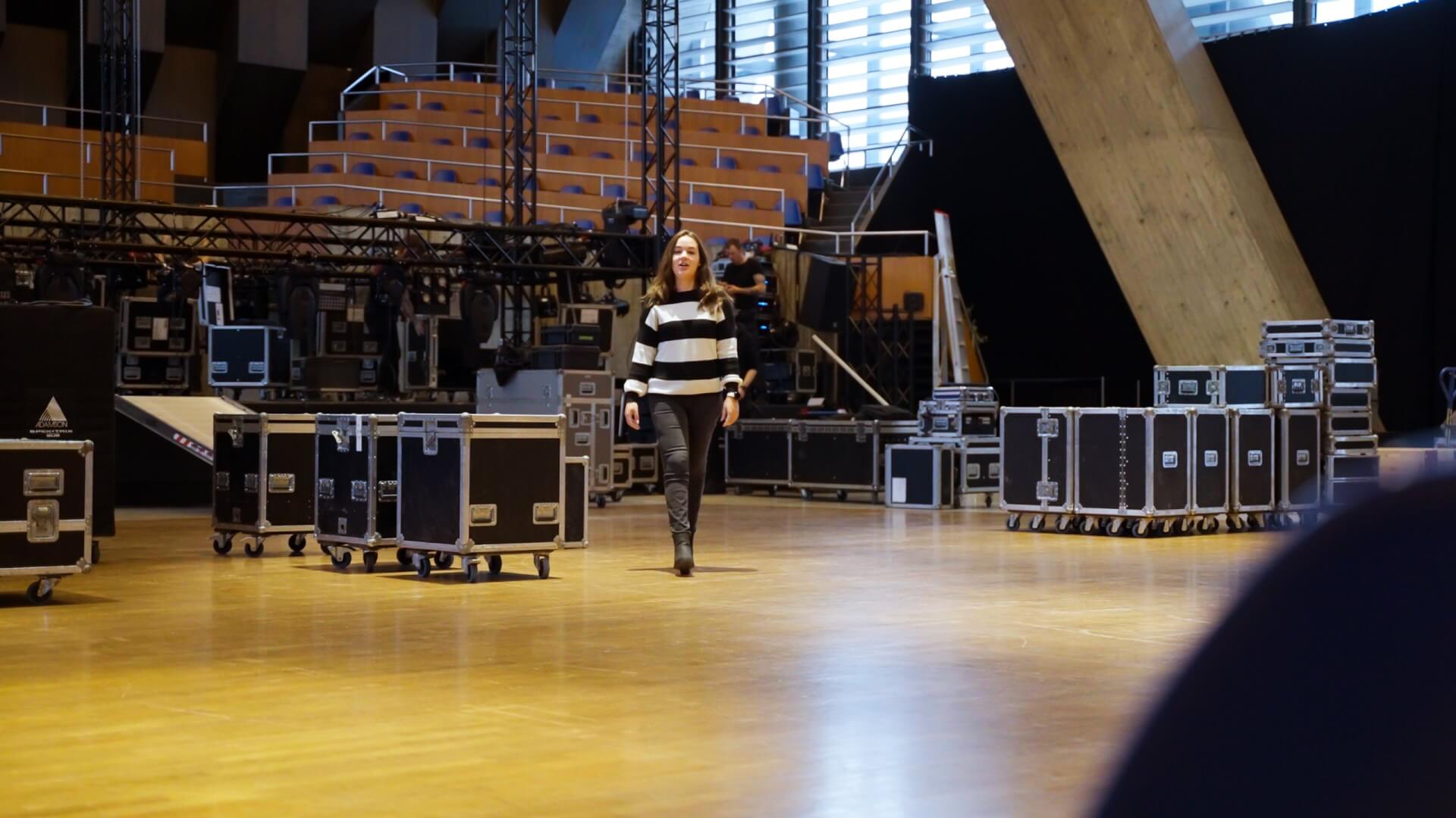 Eine Frau geht über den Boden eines geräumigen Auditoriums, in dem sich im Hintergrund Technikkästen und Bühnenaufbauten befinden, die den Alltag vom Leiter Eismeister in der Tissot Arena widerspiegeln.