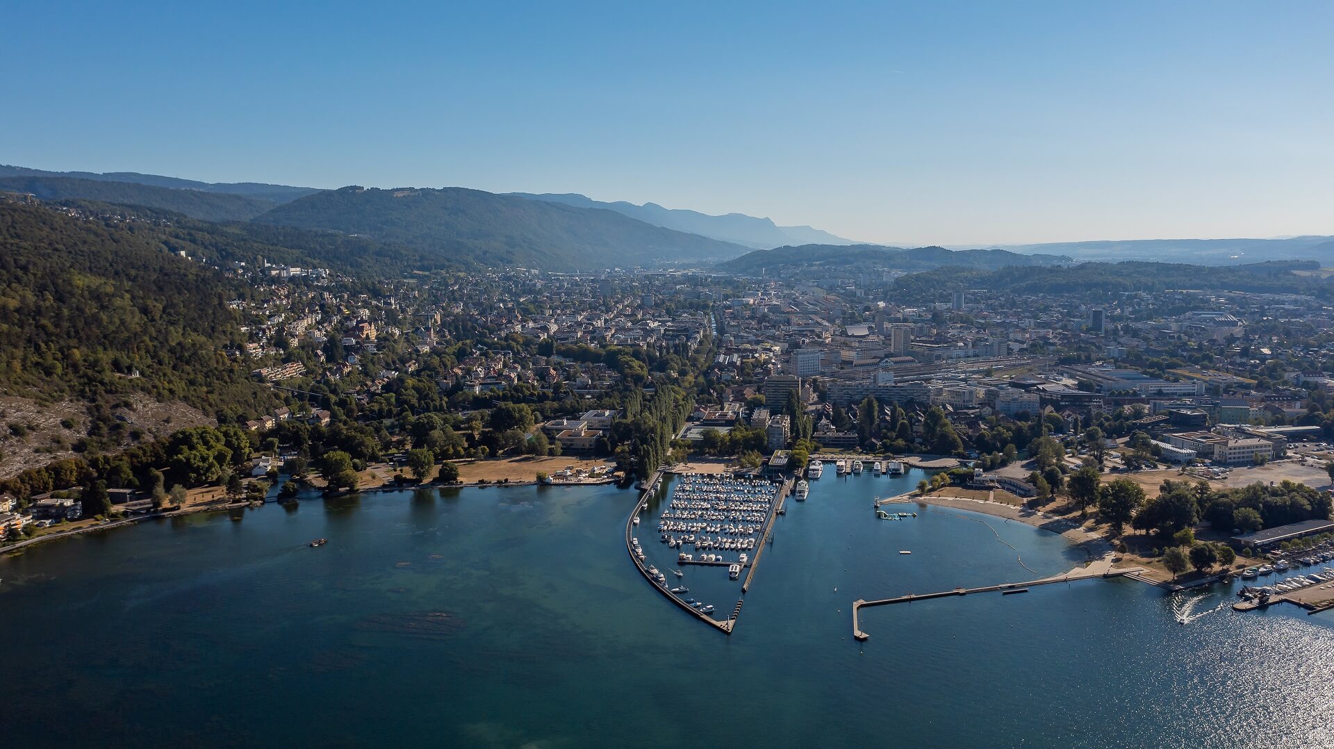 Luftaufnahme eines Jachthafens mit angedockten Booten, umgeben von einer Stadt und Bergen im Hintergrund unter einem klaren blauen Himmel.