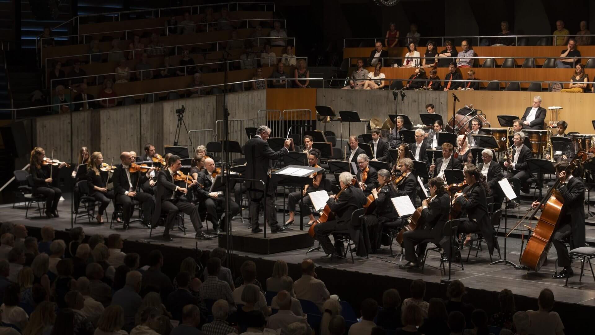 Ein Sinfonieorchester spielt unter der Leitung eines Dirigenten auf der Bühne, wobei die Musiker verschiedene Instrumente spielen und das Publikum davor sitzt.