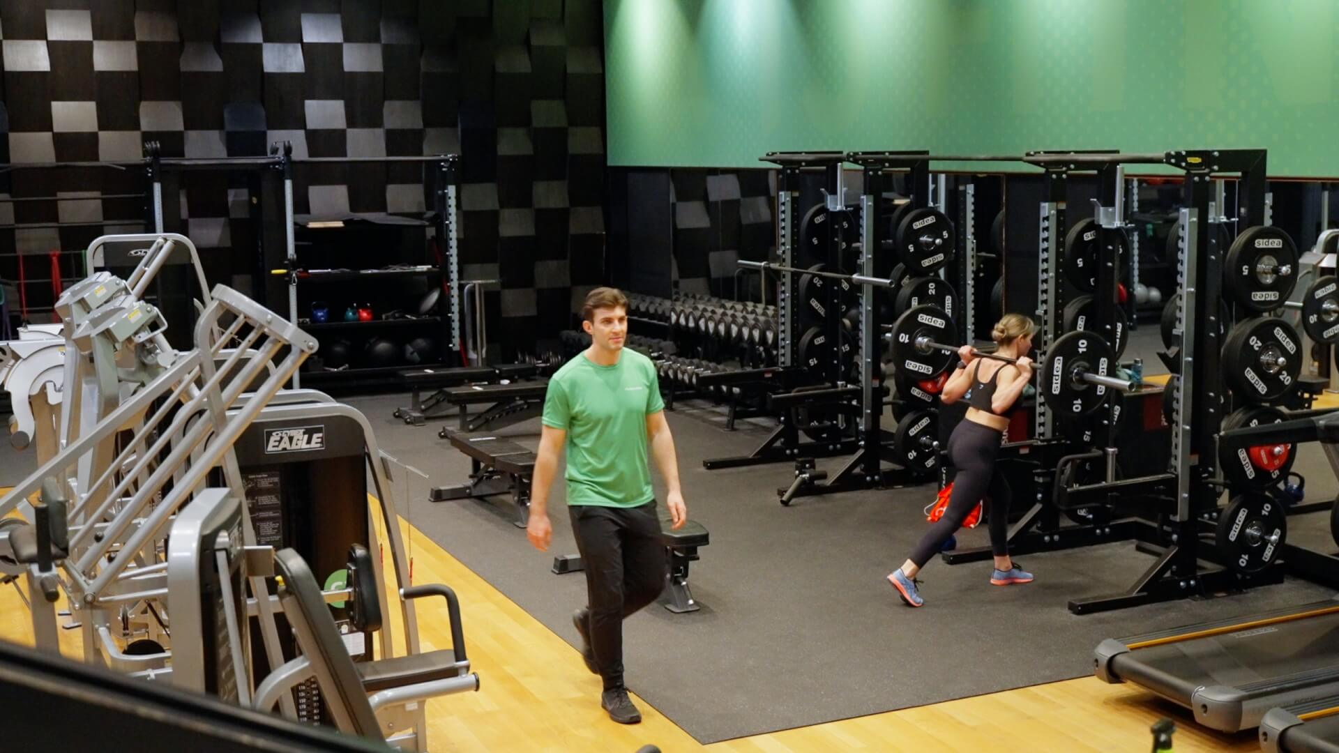 Ein Mann in einem grünen Hemd geht durch ein Fitnessstudio, während eine Frau in Sportkleidung im Hintergrund ein Squat Rack benutzt.