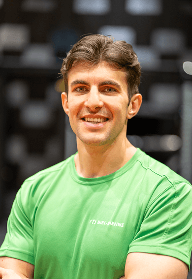 Ein Mann mit kurzen braunen Haaren und einem hellgrünen Hemd lächelt mit verschränkten Armen vor einem unscharfen Innenhintergrund in die Kamera.