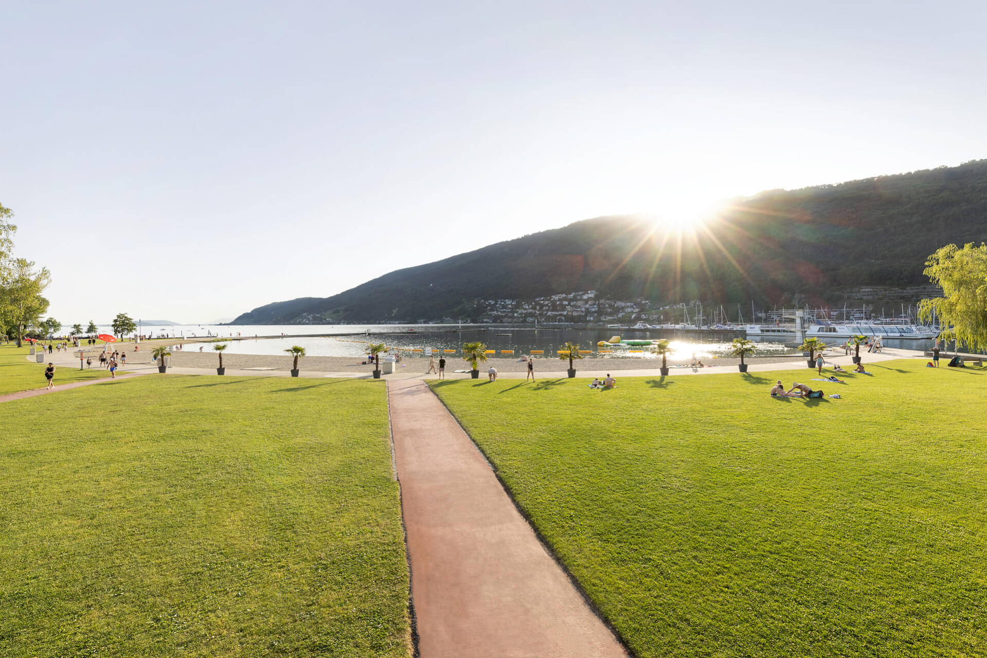 Menschen gehen spazieren und entspannen sich auf einer Wiese in der Nähe einer Uferpromenade mit kleinen Palmen, während die Sonne hinter einem Hügel untergeht. Im Hintergrund sind Boote und ein Jachthafen zu sehen.