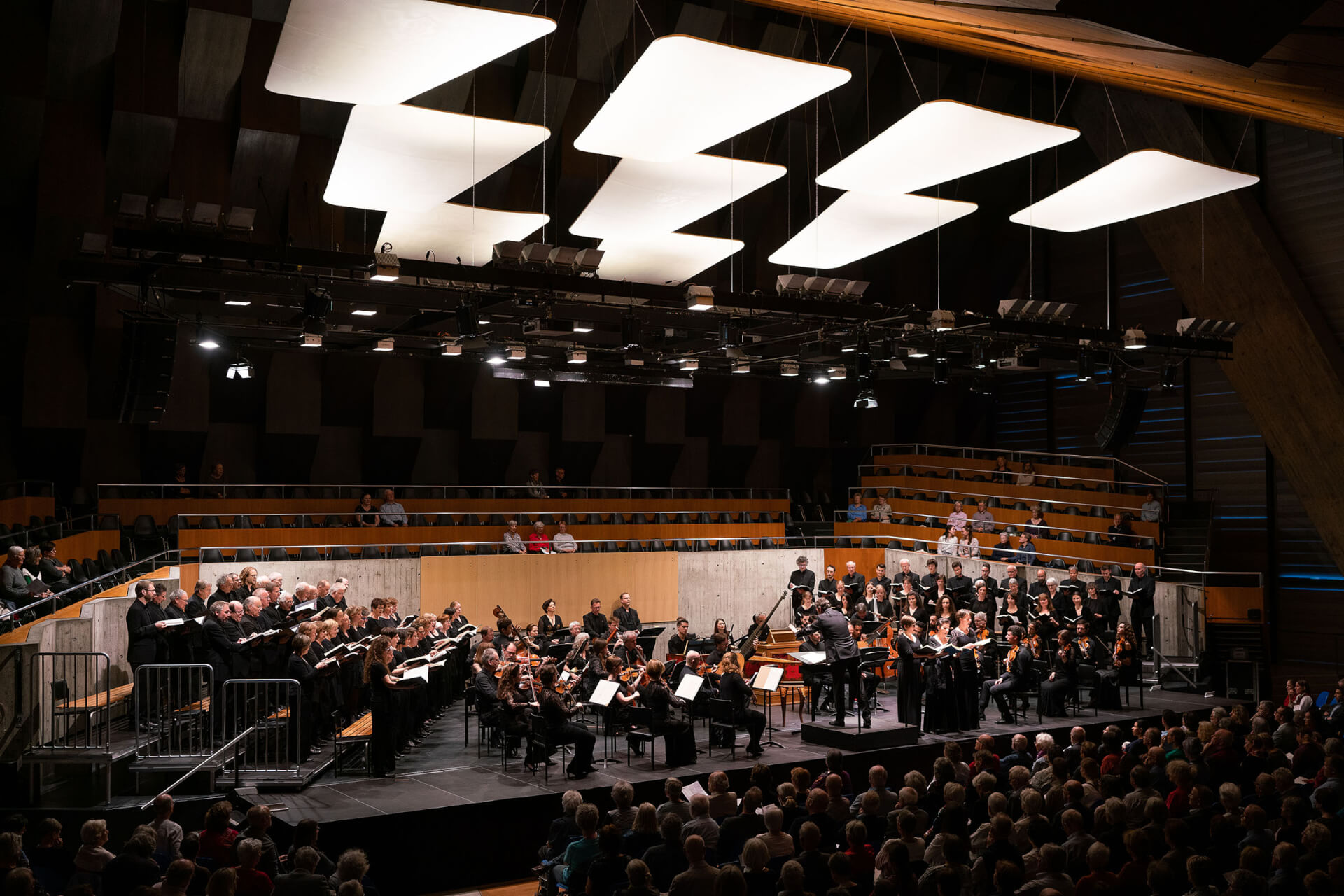 Ein Orchester und ein Chor treten auf der Bühne eines modernen Konzertsaals auf, vor dem das Publikum sitzt und über dem helle Deckenplatten angebracht sind.