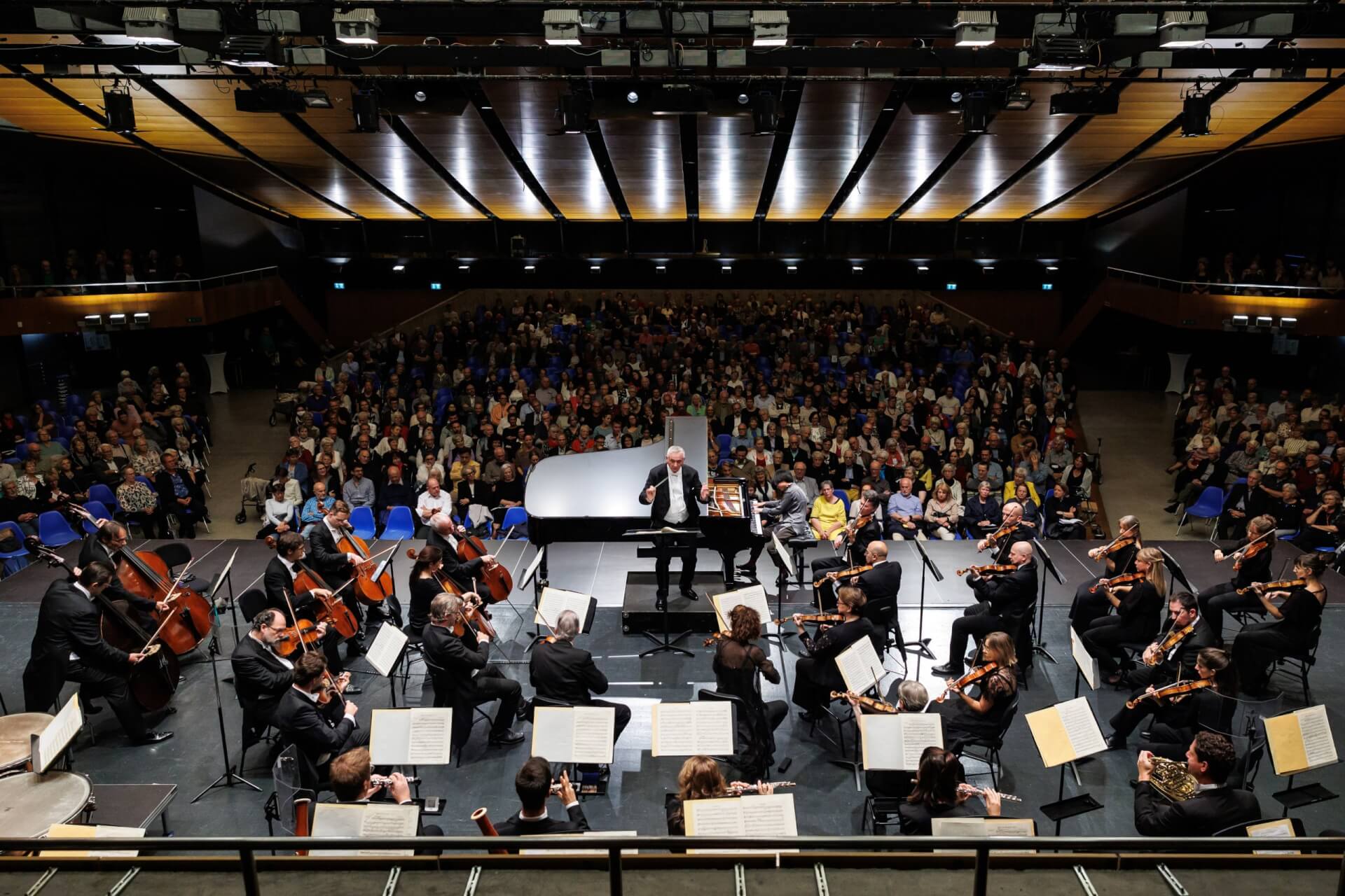 Ein Symphonieorchester spielt auf der Bühne mit einem Dirigenten, Musikern, die verschiedene Instrumente spielen, und einem großen Publikum, das im Hintergrund sitzt.