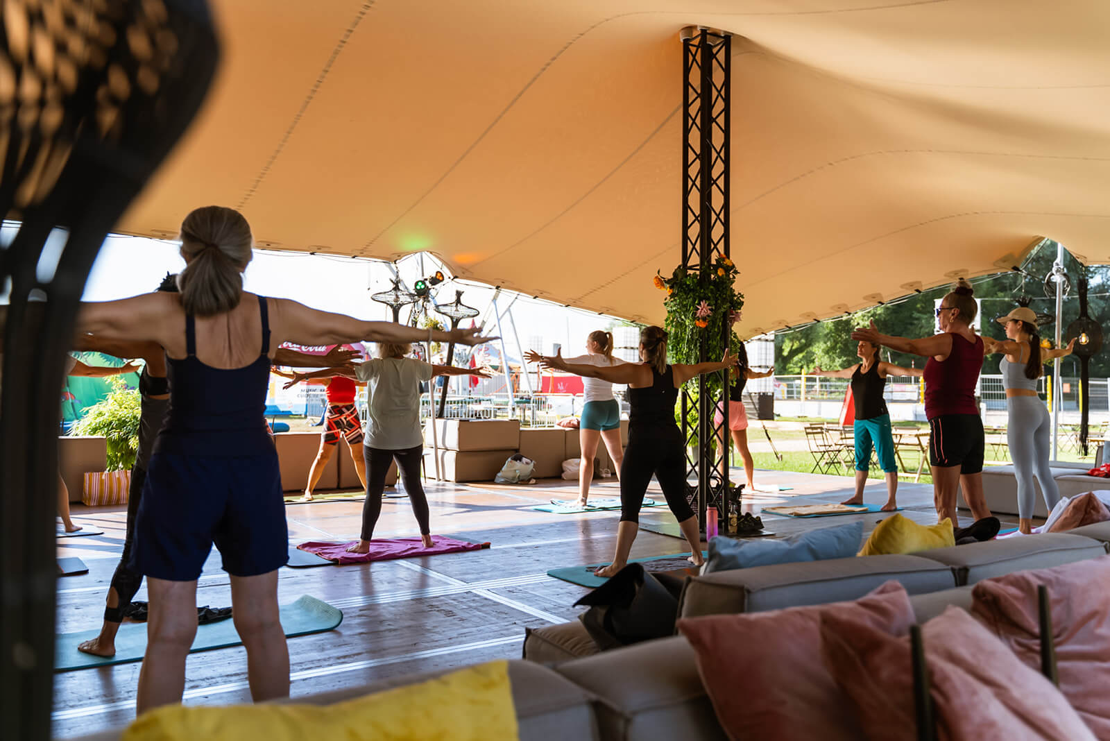 Eine Gruppe von Personen nimmt an einem Yoga- oder Fitnesskurs unter einem großen Außenzelt teil, wobei sie auf Matten stehen und die Arme zur Seite strecken.