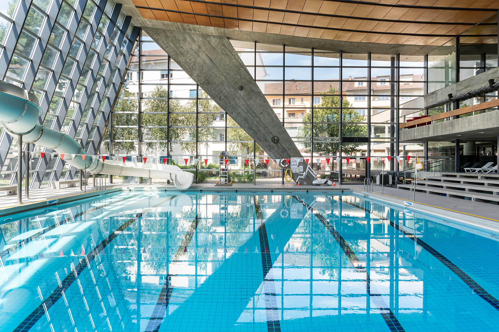 Hallenbad mit klarem Wasser, Wasserrutschen auf der linken Seite, grosse Fenster und Liegestühle auf der rechten Seite. Draussen sind städtische Gebäude und Bäume zu sehen.