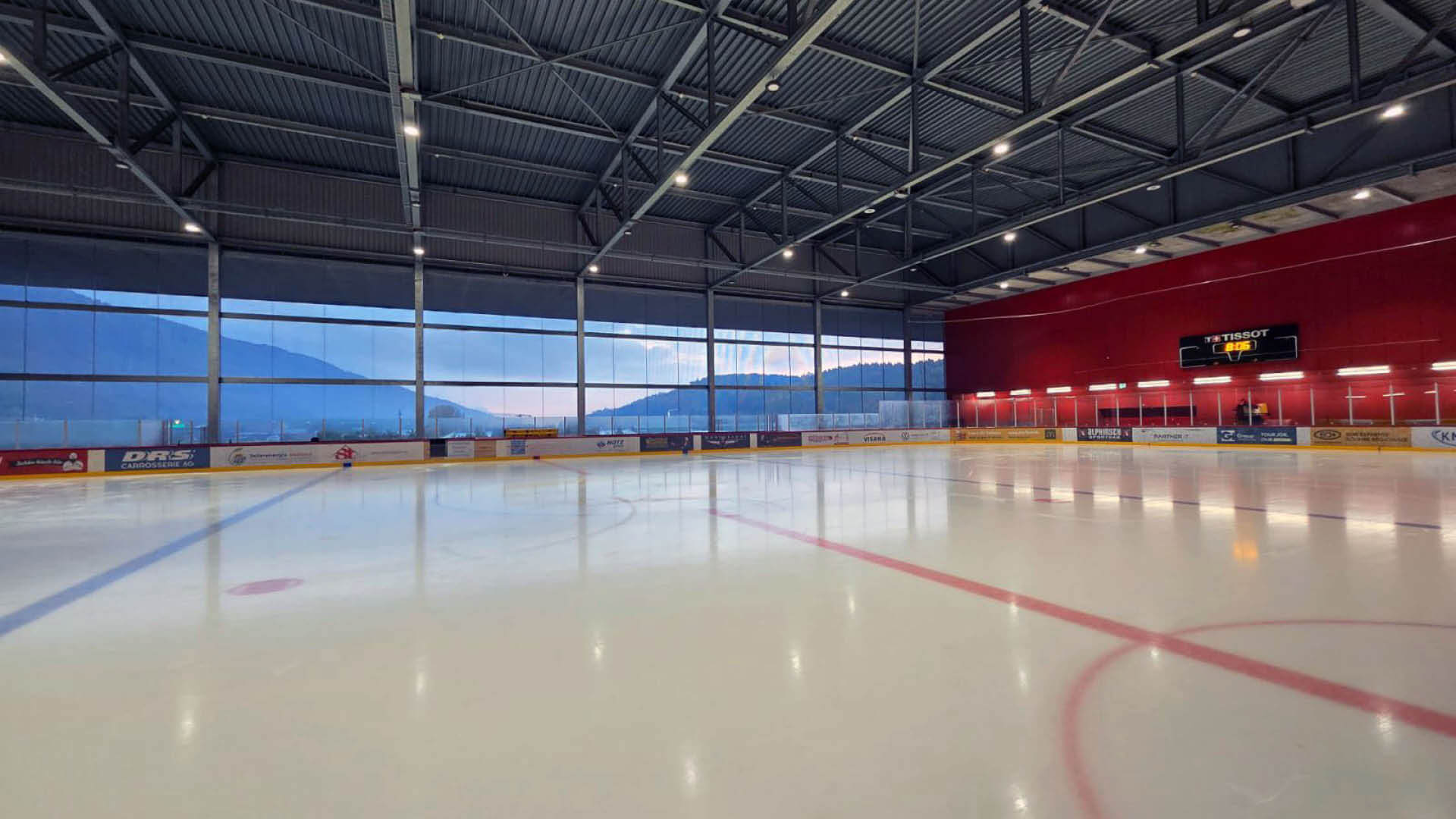 Eishockeyhalle mit glatter Eisfläche, leerer Arena, Anzeigetafel an der rechten Wand und großen Fenstern mit Bergen im Hintergrund.