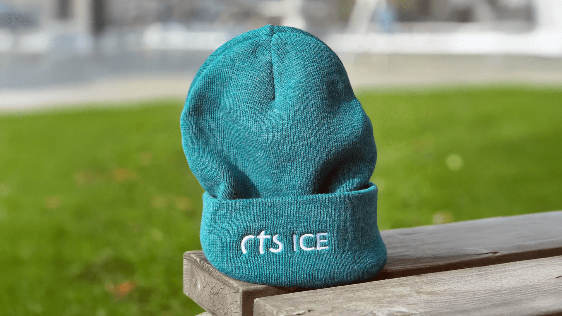 Eine blaugrüne Mütze mit dem weiß aufgestickten Schriftzug rTS ICE sitzt auf einer Holzbank im Freien, wobei Gras und ein verschwommener Hintergrund zu sehen sind.