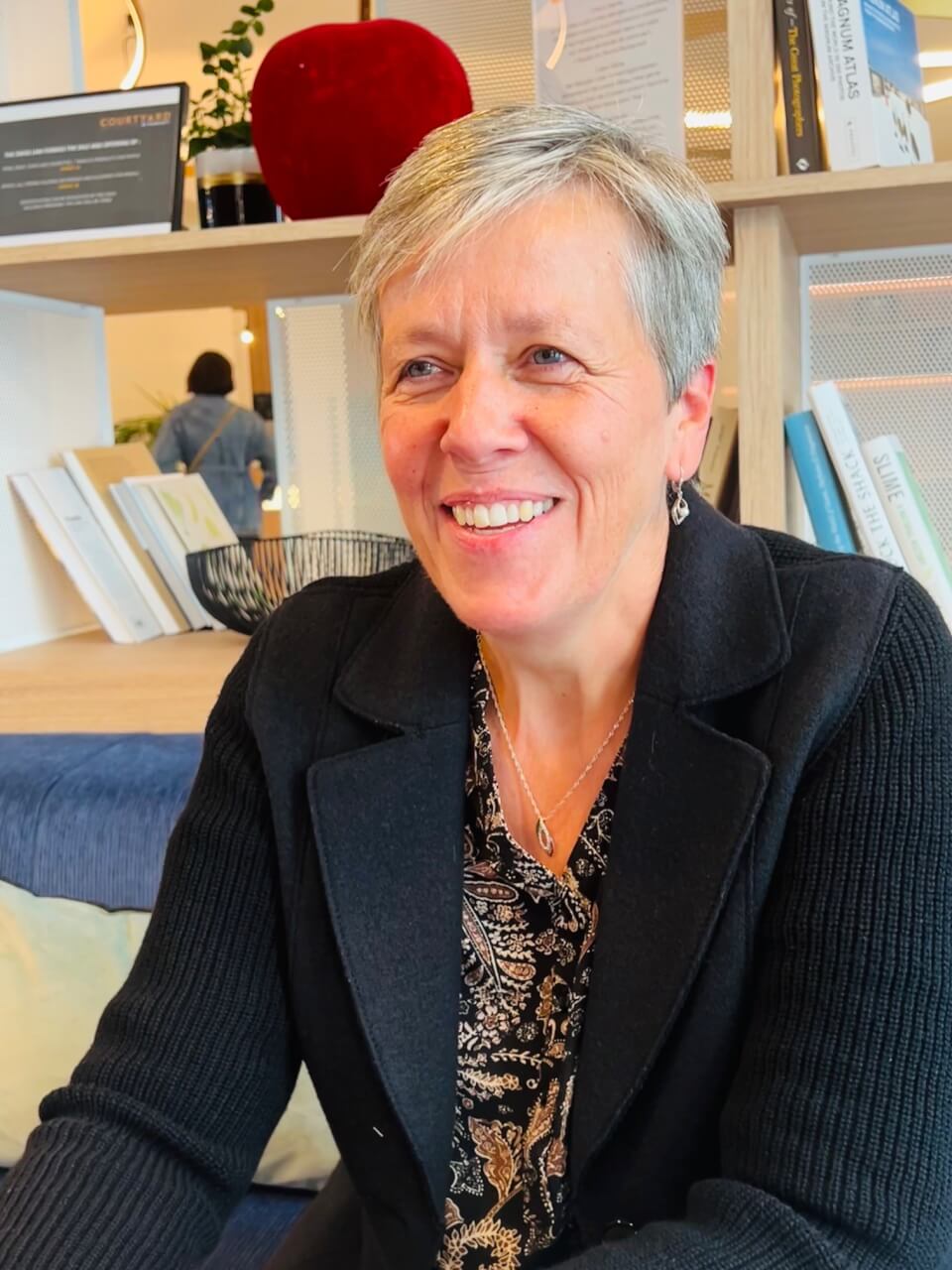 Une femme aux cheveux gris courts, vêtue d'un blazer noir et d'un chemisier à motifs, est assise et sourit dans une pièce moderne avec des étagères de livres et de décoration derrière elle.