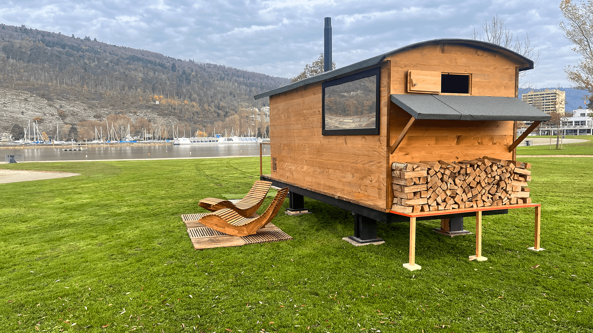 Eine hölzerne Saunahütte auf Stelzen mit gestapeltem Brennholz darunter, zwei hölzerne Liegestühle daneben, auf einer Wiese in der Nähe eines Gewässers und von Bergen.