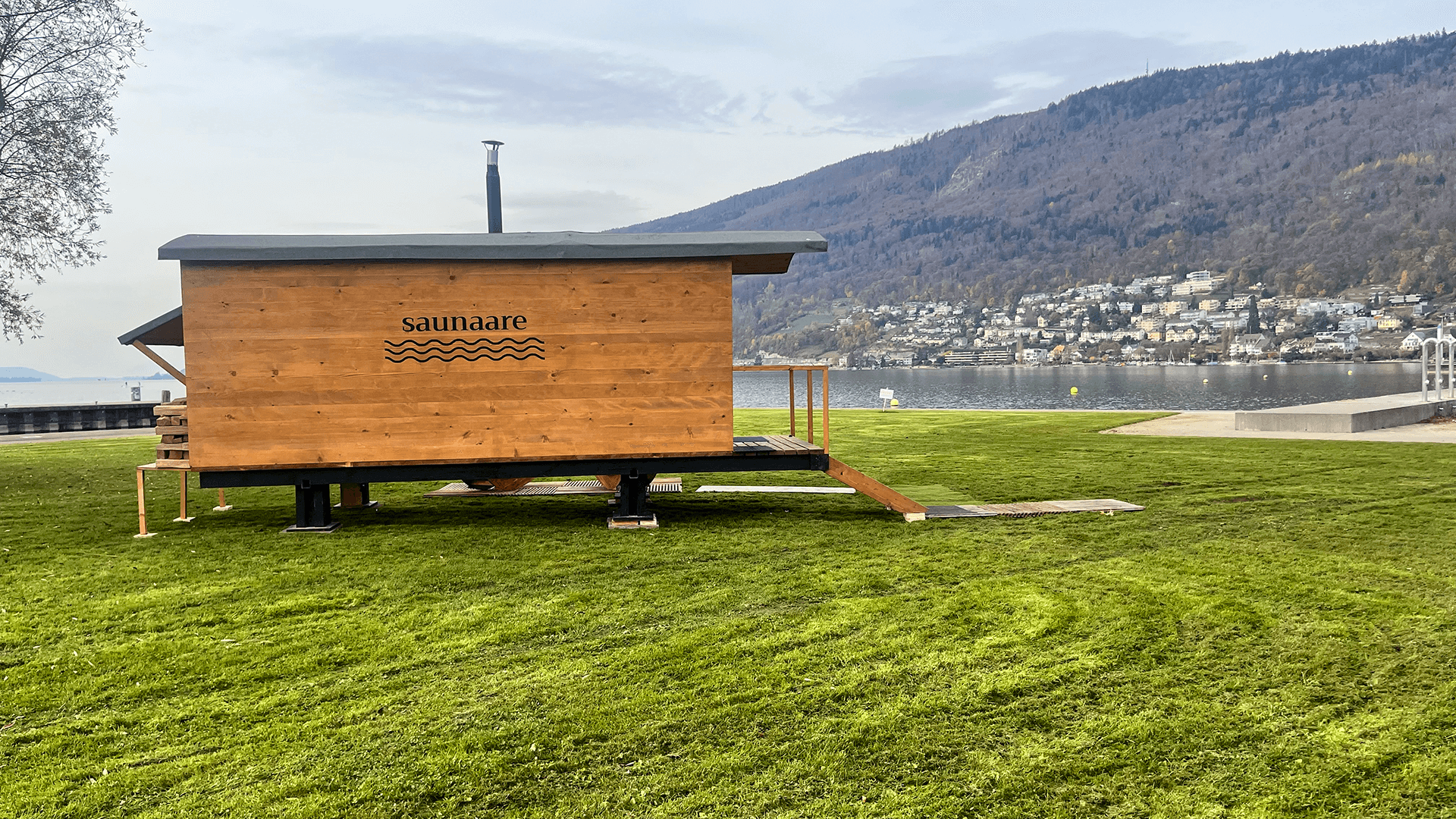 Ein hölzernes Saunagebäude mit der Bezeichnung saunaare steht auf einer Wiese in der Nähe eines Sees, mit Hügeln und verstreuten Häusern im Hintergrund.
