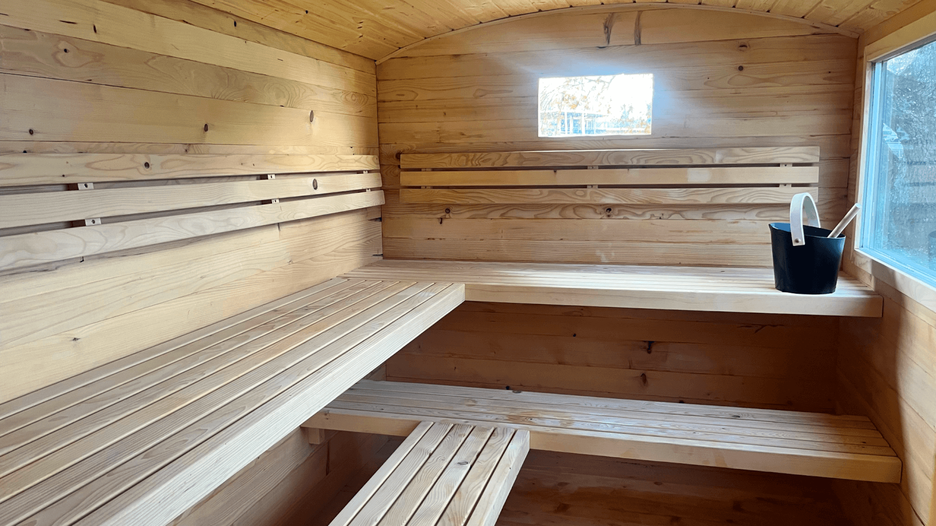 Innenraum einer Holzsauna mit gestuften Bänken, einem kleinen Fenster und einem schwarzen Eimer mit einer Schöpfkelle auf der rechten Bank.