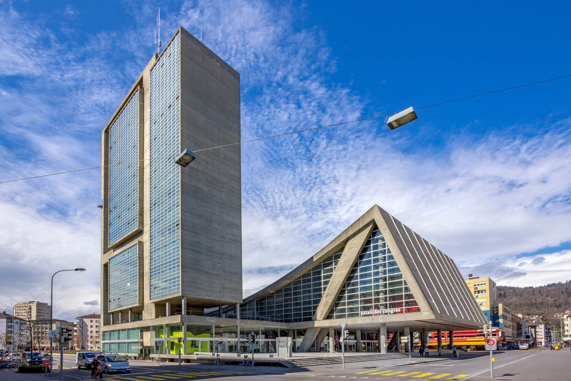 Ein modernes Gebäude mit einem hohen rechteckigen Turm und einem dreieckigen Glasaufbau steht an einer belebten Stadtkreuzung unter einem blauen Himmel mit vereinzelten Wolken.