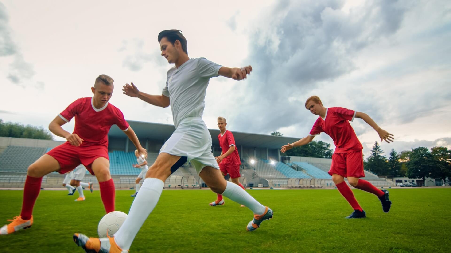 Vier männliche Fußballspieler kämpfen in einem Stadion unter bewölktem Himmel auf einem grünen Feld um den Ball, drei davon in roten und einer in weißen Trikots.