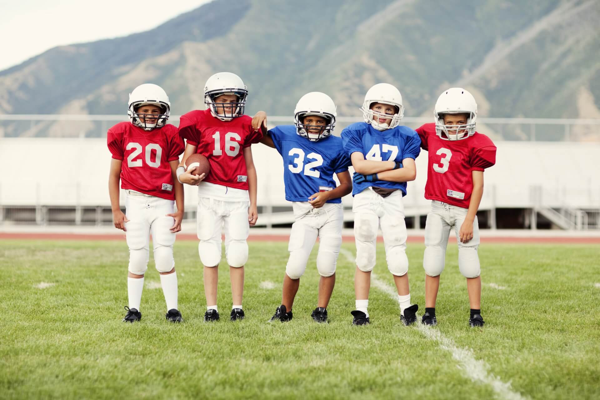 Fünf kleine Kinder in Fußballtrikots und Helmen stehen auf einer Wiese und posieren für ein Gruppenfoto vor einer Bergkulisse.