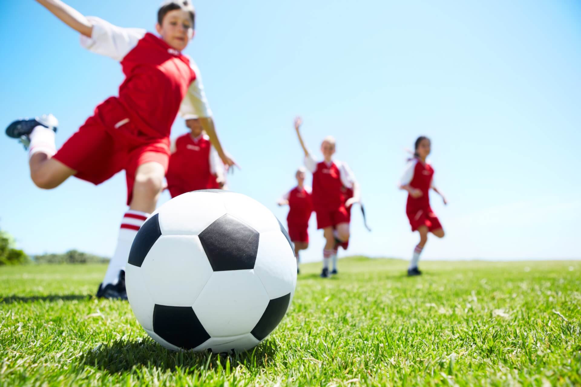 Kinder in roten Uniformen spielen draußen auf einer Wiese unter blauem Himmel Fußball, wobei ein Kind im Vordergrund einen Fußball kickt.