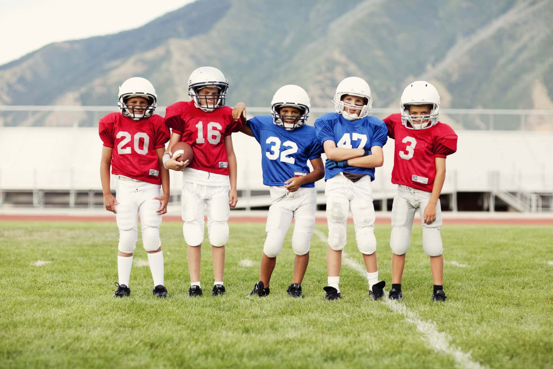 Cinq jeunes garçons portant des uniformes et des casques de football se tiennent sur un terrain herbeux avec des montagnes en arrière-plan. Trois d'entre eux portent des maillots rouges et deux des maillots bleus.