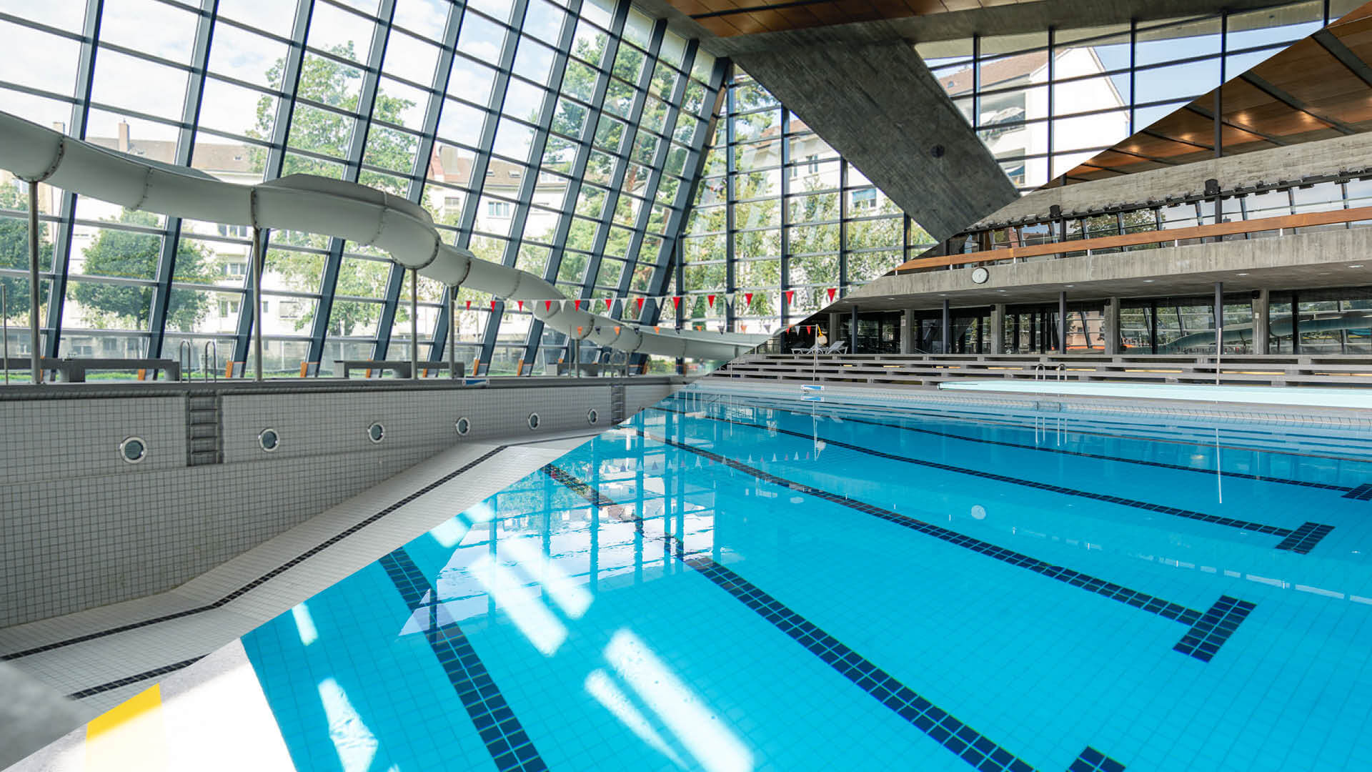 Piscine couverte aux eaux d'un bleu limpide, dotée de plusieurs couloirs, d'un toboggan aquatique et de grandes baies vitrées inclinées laissant entrer la lumière naturelle.