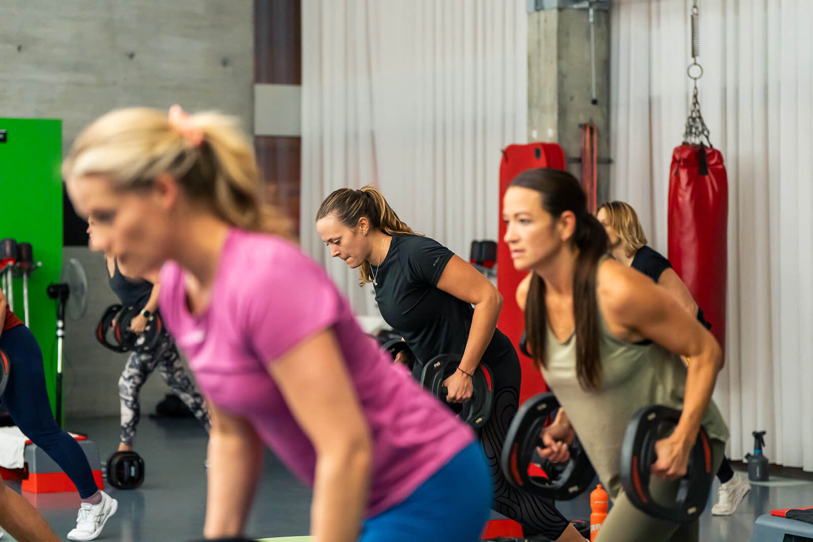 Eine Gruppe von Frauen trainiert in einem Fitnessstudio und stemmt in einem Fitnesskurs Hanteln. Im Hintergrund sind Boxausrüstung und Trainingsbänke zu sehen.