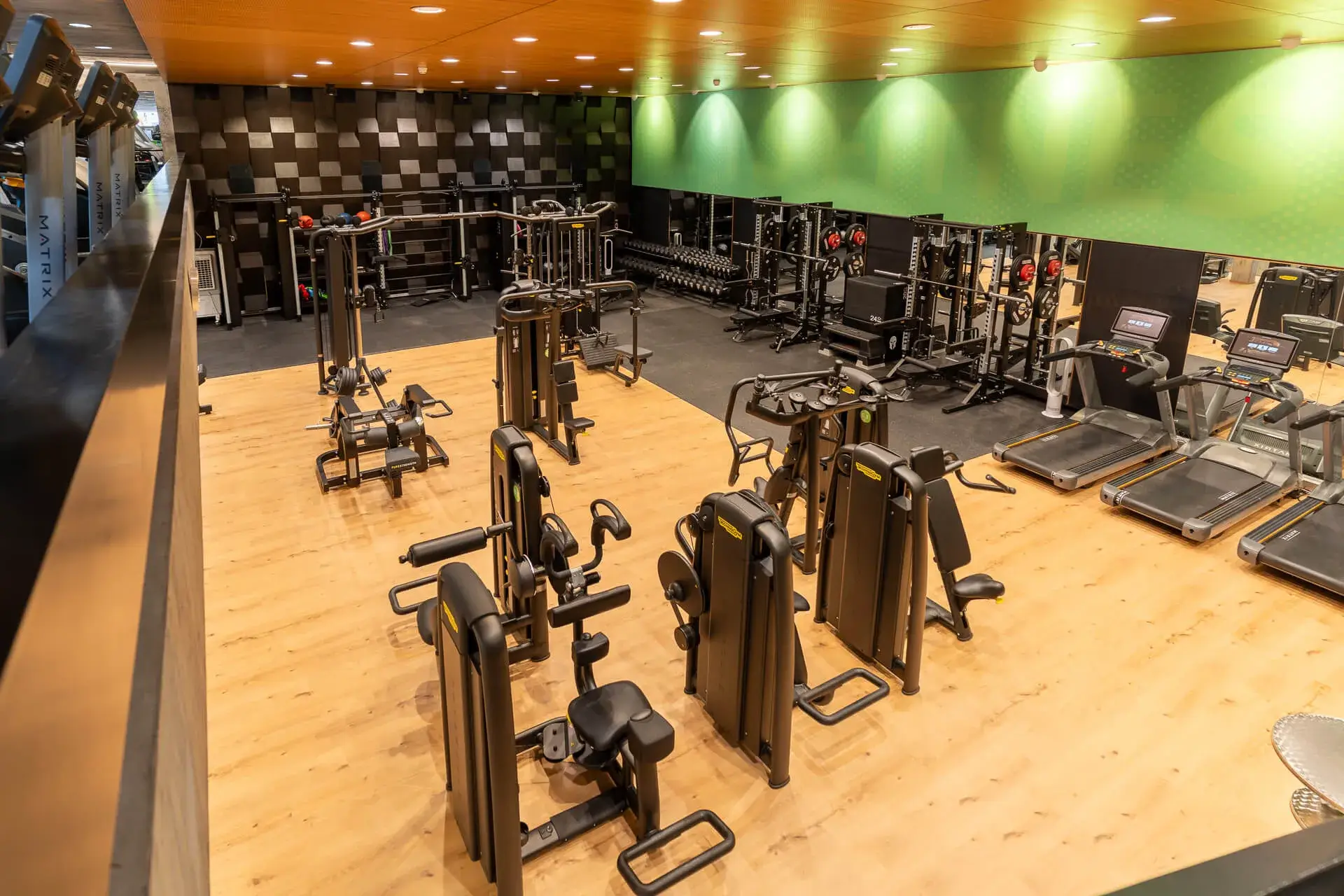 Ein modernes Fitnessstudio mit verschiedenen Trainingsgeräten, Laufbändern und Hanteln, die auf einem Holzboden unter hellen Deckenleuchten angeordnet sind.
