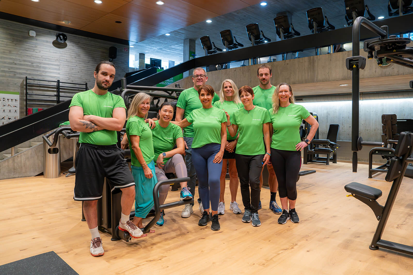 Neun Personen in einheitlichen grünen T-Shirts stehen gemeinsam in einer Sporthalle und posieren zwischen den Trainingsgeräten auf dem Holzboden für ein Gruppenfoto.
