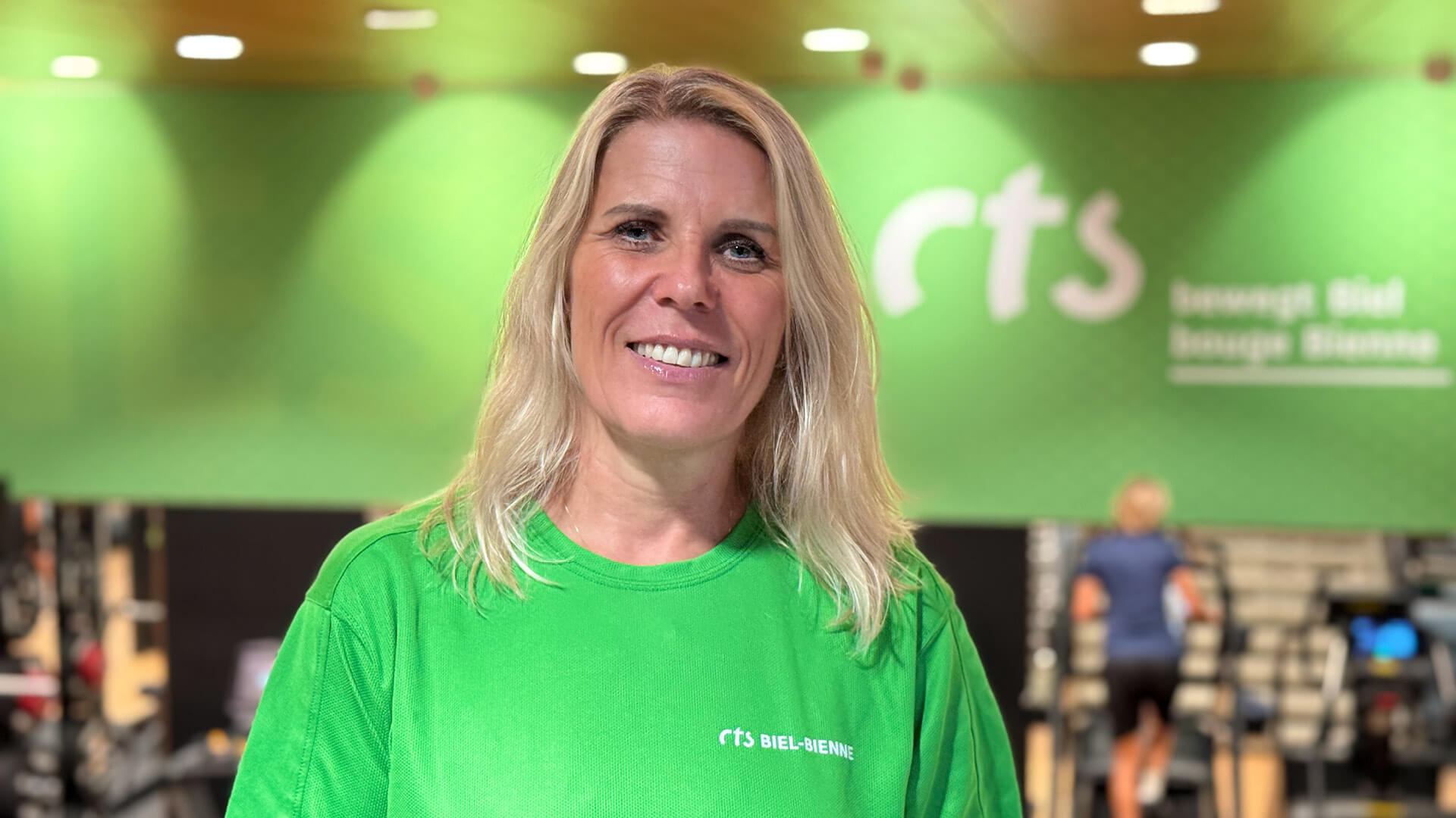 Une femme souriante aux cheveux blonds, vêtue d'un t-shirt vert « RTS Biel-Bienne », se tient à l'intérieur devant un décor de salle de sport.