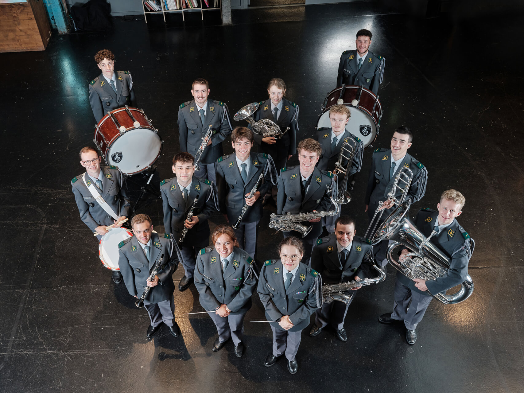 Quatorze personnes vêtues d’uniformes gris identiques posent en demi-cercle, tenant divers instruments à vent et à percussion, debout sur un sol sombre et brillant.