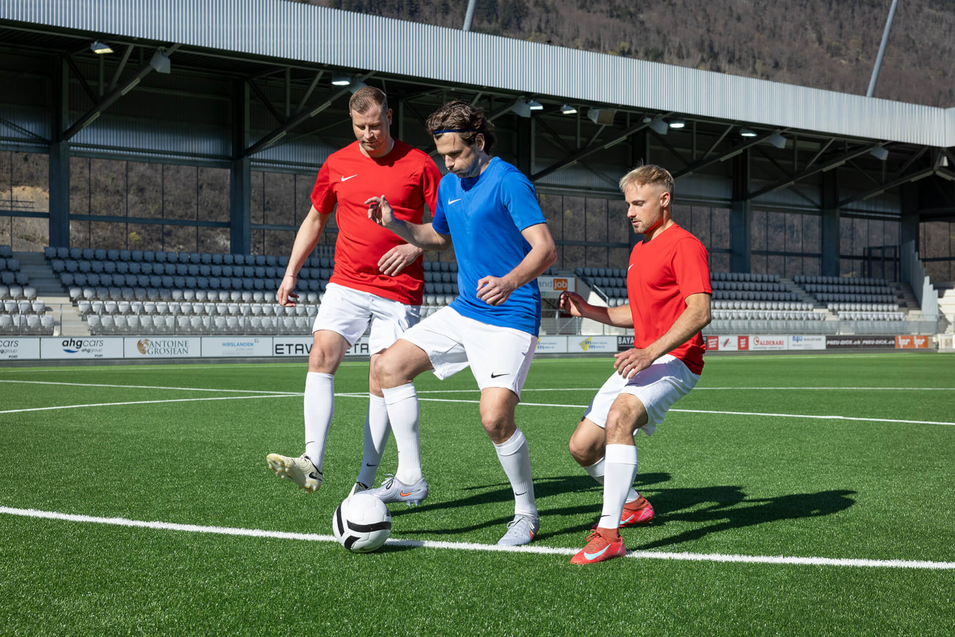 Drei Fußballspieler auf dem Platz – zwei in roten Trikots und einer in einem blauen – kämpfen während eines Spiels um den Besitz eines schwarz-weißen Fußballs.