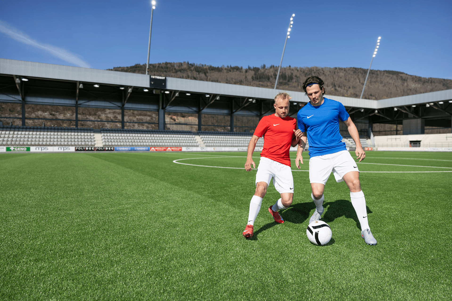 Zwei Fußballspieler in roten und blauen Trikots kämpfen auf einem grünen Stadionrasen unter strahlend blauem Himmel um den Ball.