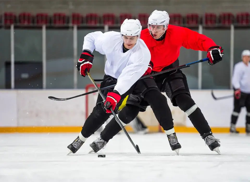 Zwei Eishockeyspieler in weißen und roten Trikots kämpfen auf einer Eishalle um den Puck, während im Hintergrund ihre Mannschaftskameraden und leere Sitzplätze zu sehen sind.